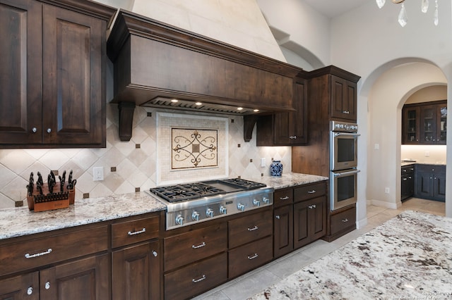 kitchen with light tile patterned floors, dark brown cabinets, backsplash, appliances with stainless steel finishes, and custom range hood