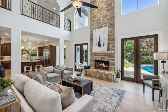 living room featuring a stone fireplace, ceiling fan with notable chandelier, a towering ceiling, french doors, and light hardwood / wood-style floors