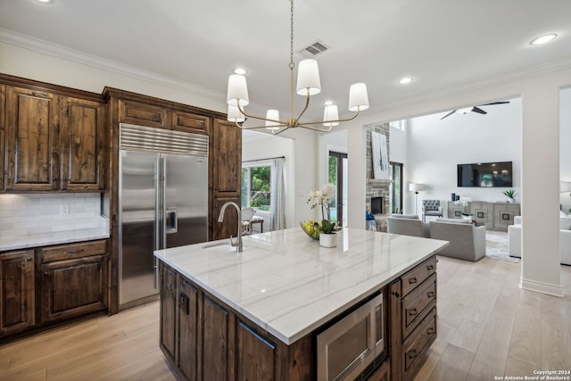 kitchen with a kitchen island with sink, light wood-type flooring, built in appliances, pendant lighting, and sink