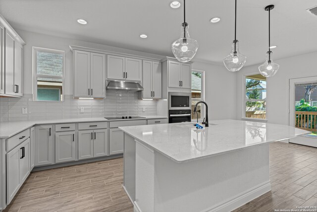 kitchen with stainless steel appliances, light stone countertops, a center island with sink, and hanging light fixtures