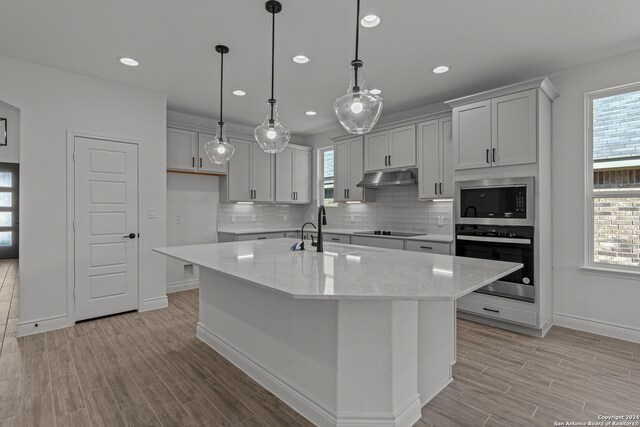 kitchen featuring a center island with sink, hanging light fixtures, appliances with stainless steel finishes, light stone countertops, and light hardwood / wood-style flooring