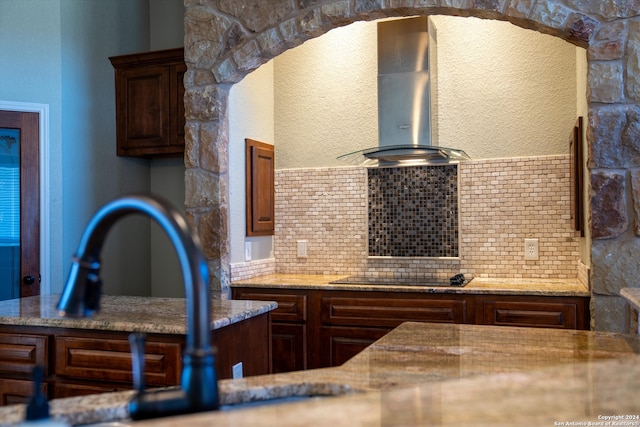 kitchen with wall chimney range hood, backsplash, stone countertops, black electric cooktop, and dark brown cabinetry