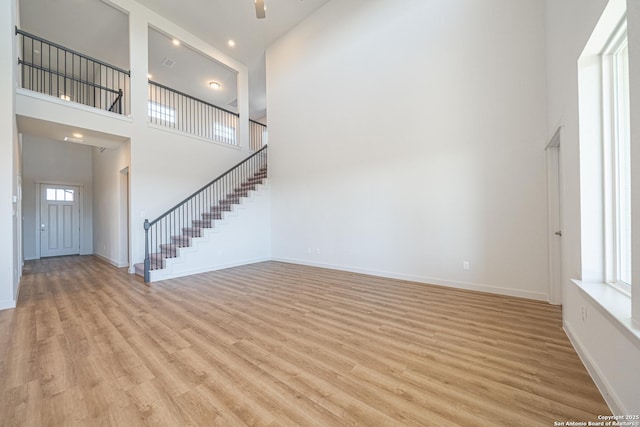 unfurnished living room featuring a towering ceiling, light wood-style flooring, stairway, and baseboards