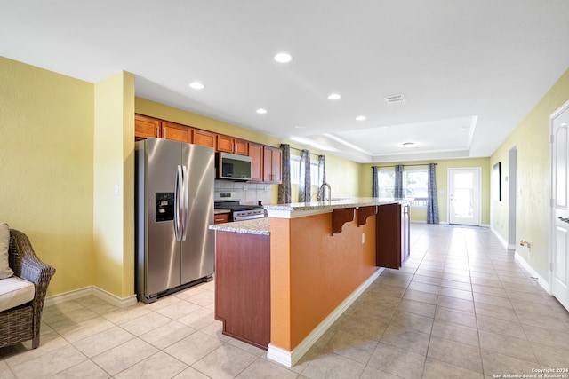 kitchen featuring appliances with stainless steel finishes, a kitchen bar, a tray ceiling, light stone counters, and a center island with sink