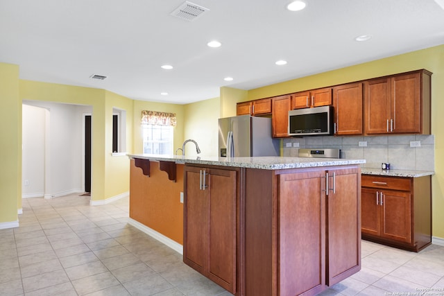 kitchen with tasteful backsplash, light stone countertops, appliances with stainless steel finishes, light tile patterned floors, and a center island with sink