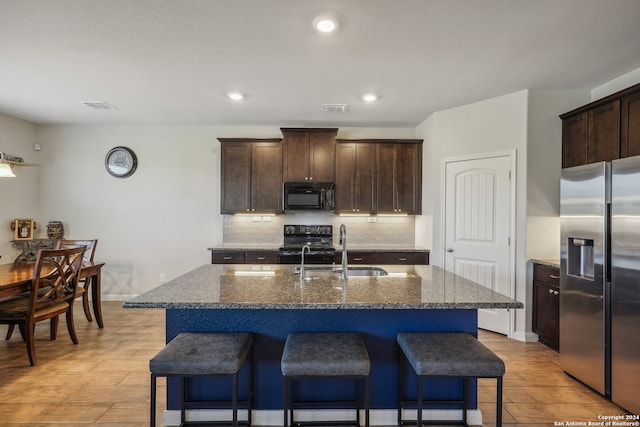 kitchen featuring an island with sink, dark stone counters, light wood-type flooring, black appliances, and sink