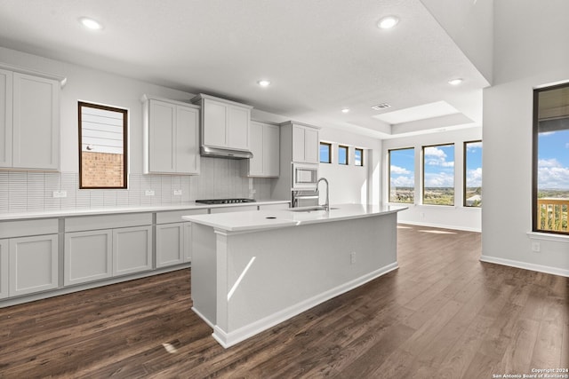 kitchen with sink, dark wood-type flooring, decorative backsplash, a center island with sink, and white microwave