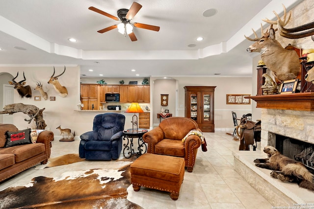 living room featuring a stone fireplace, ornamental molding, a tray ceiling, and ceiling fan