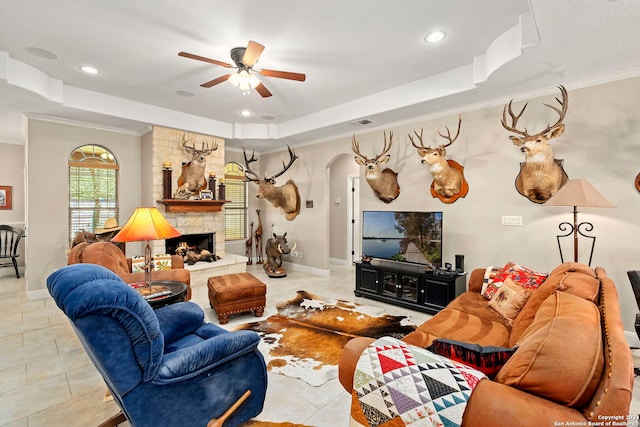 living room with a stone fireplace, a raised ceiling, ceiling fan, crown molding, and light tile patterned floors