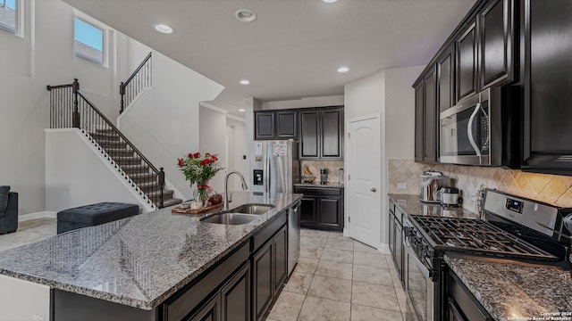 kitchen featuring a kitchen island with sink, backsplash, dark stone counters, sink, and appliances with stainless steel finishes