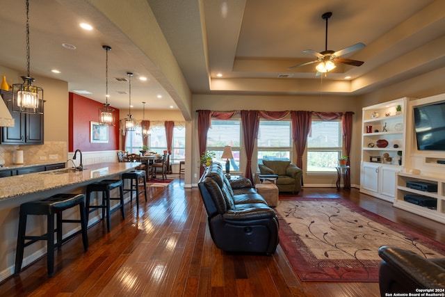 living room with sink, ceiling fan with notable chandelier, a raised ceiling, dark hardwood / wood-style floors, and built in shelves