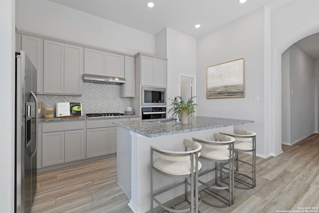kitchen featuring decorative backsplash, under cabinet range hood, light stone counters, arched walkways, and appliances with stainless steel finishes