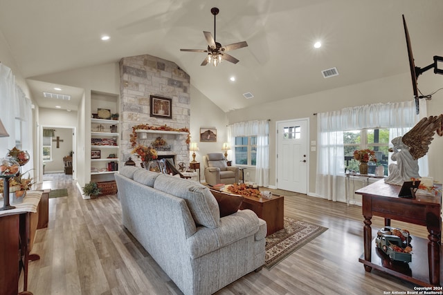 living room with light hardwood / wood-style floors, a stone fireplace, built in shelves, high vaulted ceiling, and ceiling fan