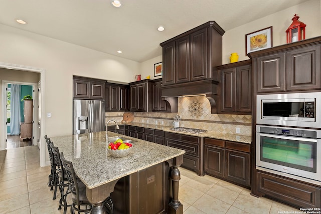 kitchen featuring a center island with sink, appliances with stainless steel finishes, light stone countertops, a kitchen bar, and sink