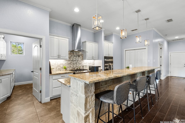 kitchen featuring wall chimney range hood, an island with sink, a kitchen breakfast bar, pendant lighting, and white cabinets