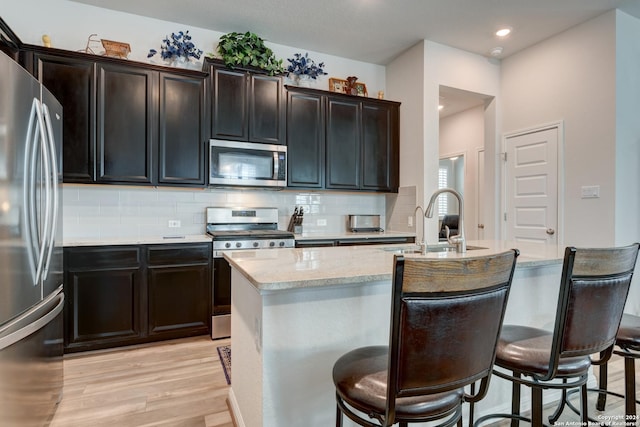 kitchen with appliances with stainless steel finishes, light wood-type flooring, backsplash, sink, and an island with sink