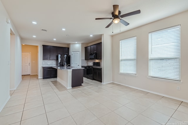 kitchen with black appliances, backsplash, ceiling fan, light stone counters, and a kitchen island with sink