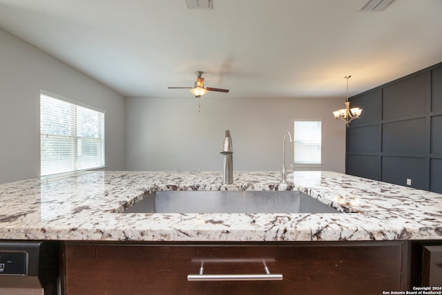 kitchen with light stone counters, sink, hanging light fixtures, and ceiling fan with notable chandelier