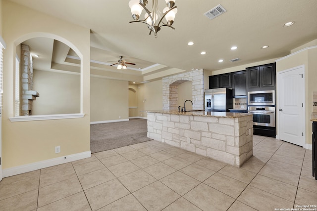 kitchen featuring a kitchen island with sink, a raised ceiling, appliances with stainless steel finishes, light stone counters, and ceiling fan with notable chandelier