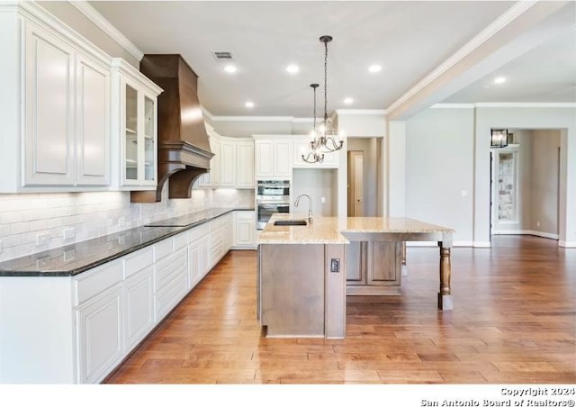 kitchen featuring white cabinets, premium range hood, a kitchen island with sink, light hardwood / wood-style flooring, and dark stone countertops