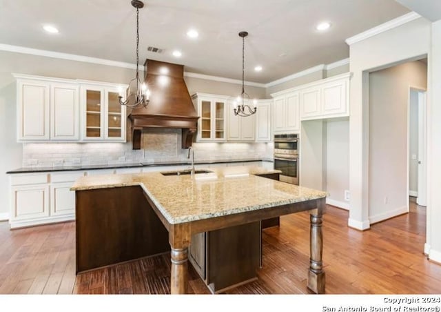 kitchen featuring custom exhaust hood, hardwood / wood-style floors, light stone countertops, and a center island with sink