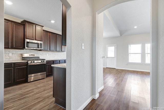 kitchen featuring dark brown cabinets, wood-type flooring, appliances with stainless steel finishes, light stone counters, and tasteful backsplash