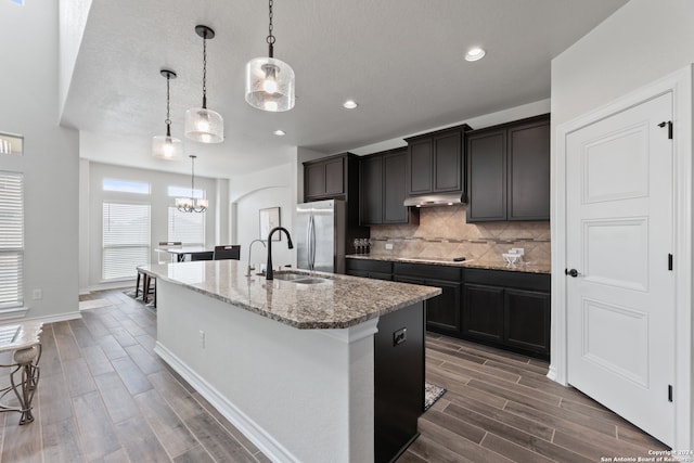 kitchen with sink, an island with sink, dark hardwood / wood-style flooring, stainless steel fridge, and decorative light fixtures