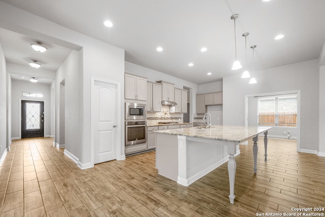 kitchen featuring appliances with stainless steel finishes, a kitchen island with sink, light stone countertops, light wood-type flooring, and pendant lighting