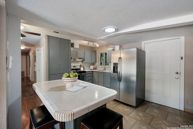 kitchen featuring tile countertops, a kitchen breakfast bar, gray cabinetry, stainless steel appliances, and a textured ceiling