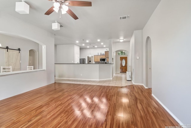 unfurnished living room featuring a barn door, ceiling fan, light wood-type flooring, visible vents, and arched walkways