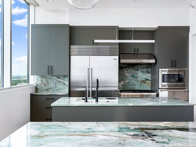 kitchen with backsplash, light stone countertops, built in appliances, gray cabinets, and sink