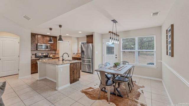 kitchen featuring an island with sink, stainless steel appliances, sink, light stone countertops, and decorative light fixtures