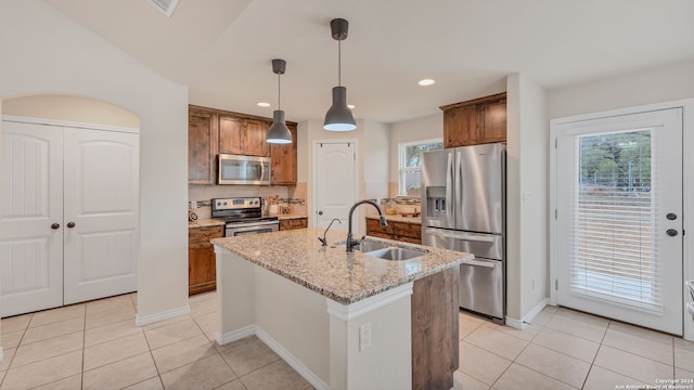 kitchen with a kitchen island with sink, light stone countertops, sink, pendant lighting, and stainless steel appliances