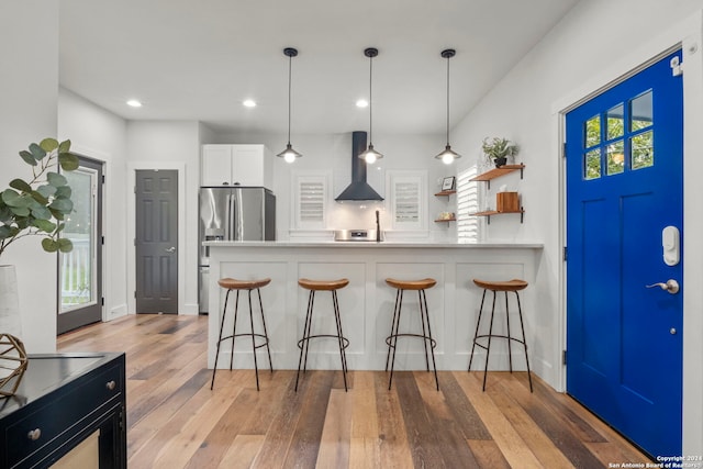 kitchen with a kitchen breakfast bar, wall chimney exhaust hood, white cabinets, stainless steel fridge, and light wood-type flooring