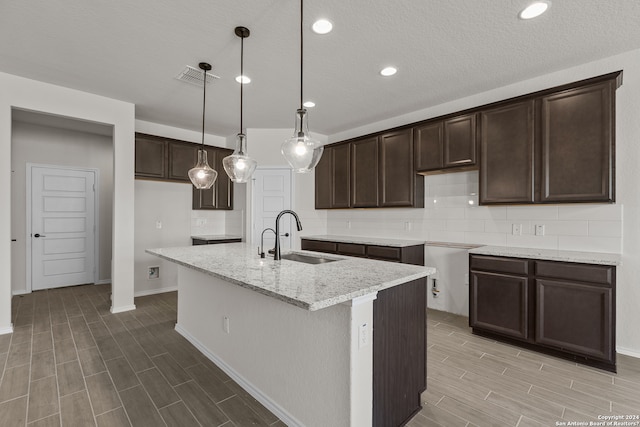 kitchen with hanging light fixtures, a center island with sink, sink, dark brown cabinetry, and light stone counters