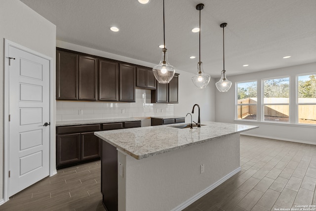 kitchen with a kitchen island with sink, light stone countertops, pendant lighting, dark brown cabinetry, and sink