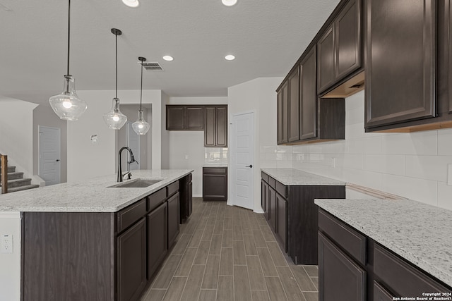 kitchen featuring sink, hanging light fixtures, dark brown cabinetry, decorative backsplash, and a kitchen island with sink