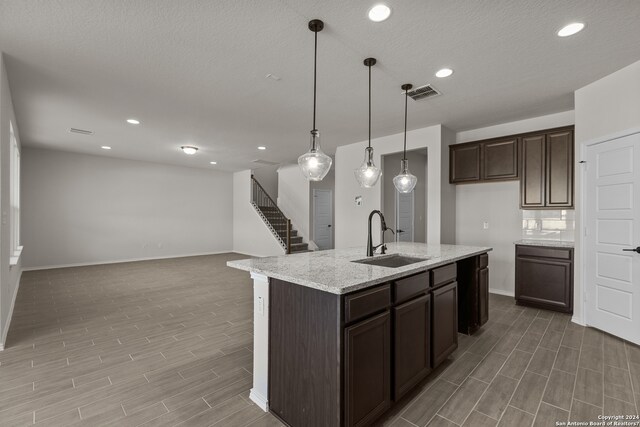 kitchen featuring hanging light fixtures, an island with sink, light stone countertops, dark brown cabinetry, and sink