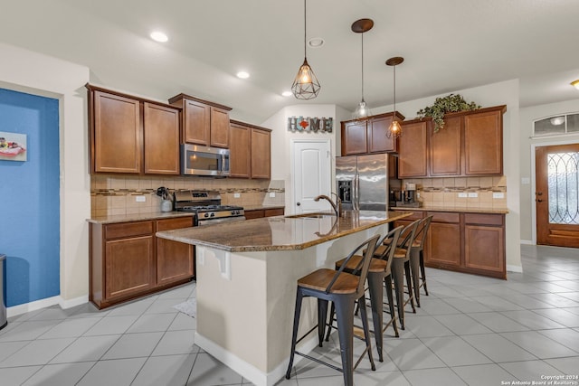 kitchen featuring a center island with sink, appliances with stainless steel finishes, dark stone counters, a kitchen bar, and decorative light fixtures