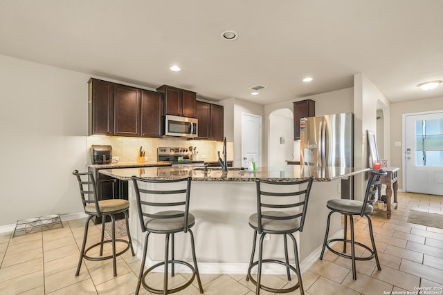 kitchen with a kitchen breakfast bar, an island with sink, stainless steel appliances, backsplash, and dark brown cabinetry