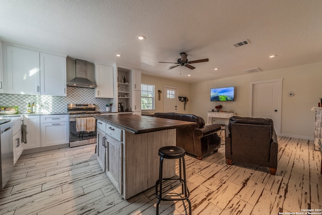 kitchen featuring a kitchen island, white cabinetry, ceiling fan, stainless steel appliances, and wall chimney exhaust hood