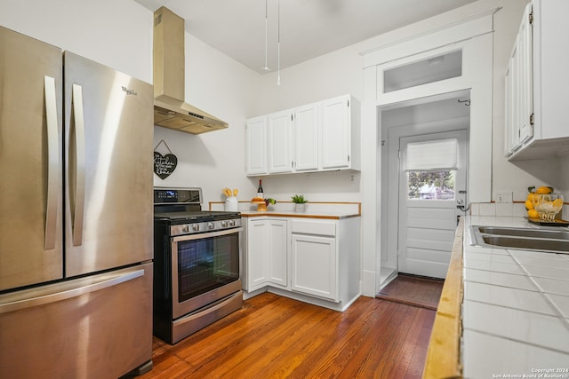 kitchen featuring wall chimney range hood, tile counters, hardwood / wood-style floors, white cabinetry, and appliances with stainless steel finishes