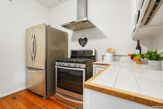 kitchen featuring tile counters, exhaust hood, stainless steel appliances, hardwood / wood-style floors, and white cabinets