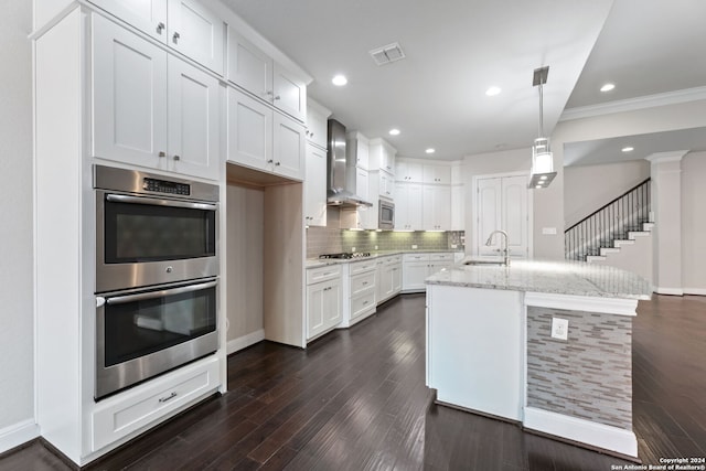 kitchen with wall chimney range hood, stainless steel appliances, white cabinets, light stone counters, and a center island with sink