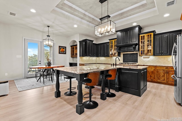 kitchen with a center island with sink, light stone countertops, light wood-type flooring, a raised ceiling, and stainless steel refrigerator