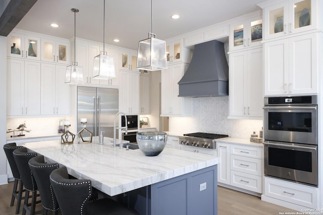 kitchen featuring white cabinets, custom range hood, an island with sink, and appliances with stainless steel finishes