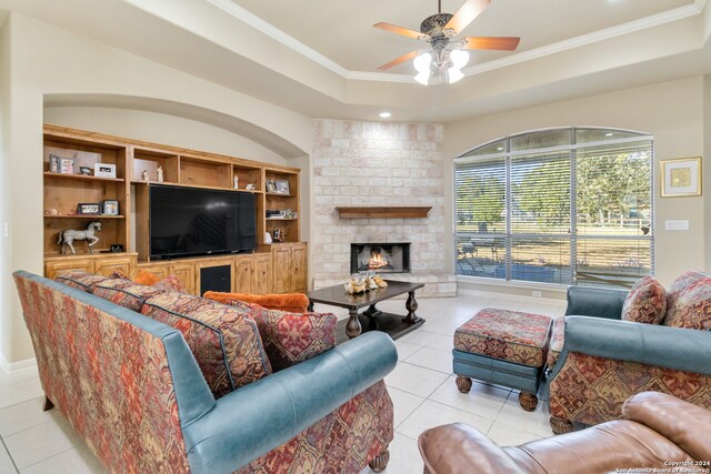 living room featuring a raised ceiling, a brick fireplace, ceiling fan, crown molding, and light tile patterned floors