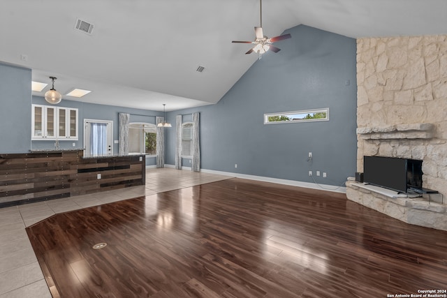 unfurnished living room featuring light hardwood / wood-style flooring, a stone fireplace, high vaulted ceiling, and ceiling fan with notable chandelier