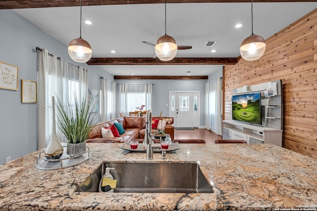 kitchen featuring light stone counters, beamed ceiling, sink, wooden walls, and decorative light fixtures