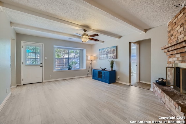 unfurnished living room featuring ceiling fan, a textured ceiling, a brick fireplace, light hardwood / wood-style flooring, and beamed ceiling